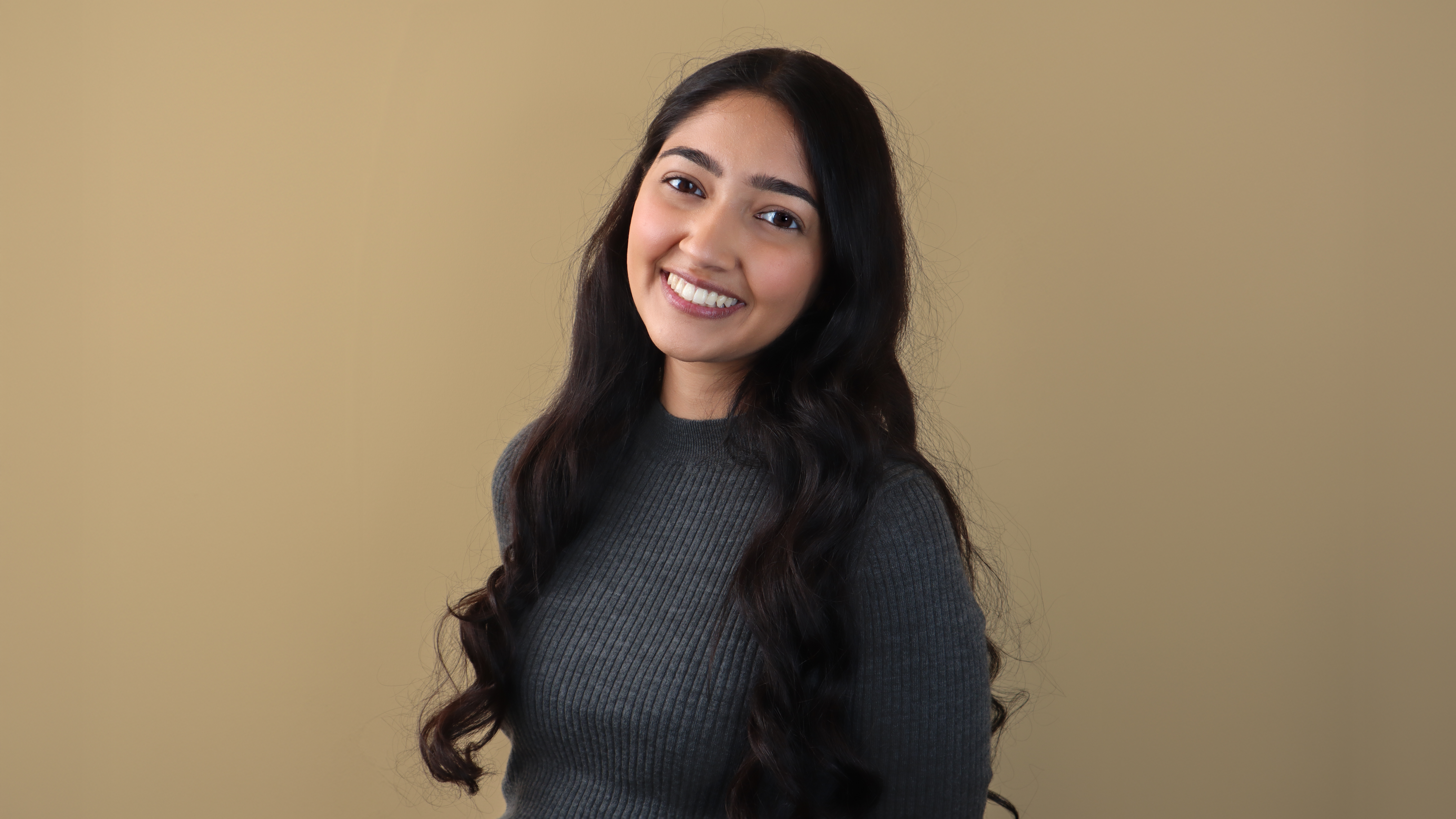 Saloni Trivedi smiles in front of a beige background wearing a gray turtleneck. Saloni Trivedi smiles in front of a beige background wearing a gray turtleneck.